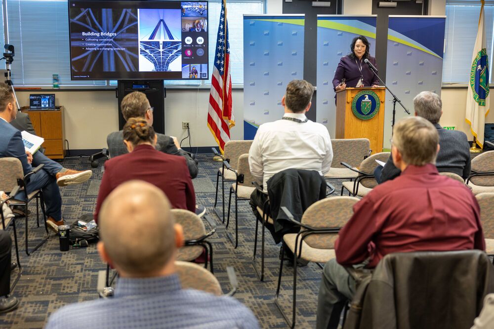 Photo show a hybrid meeting, with people in the room and a TV with virtual attendees. View from back of room with person speaking at a lectern.