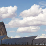 Arrays of solar panels stand in front of a prominent rock formation with scattered clouds and blue sky above.