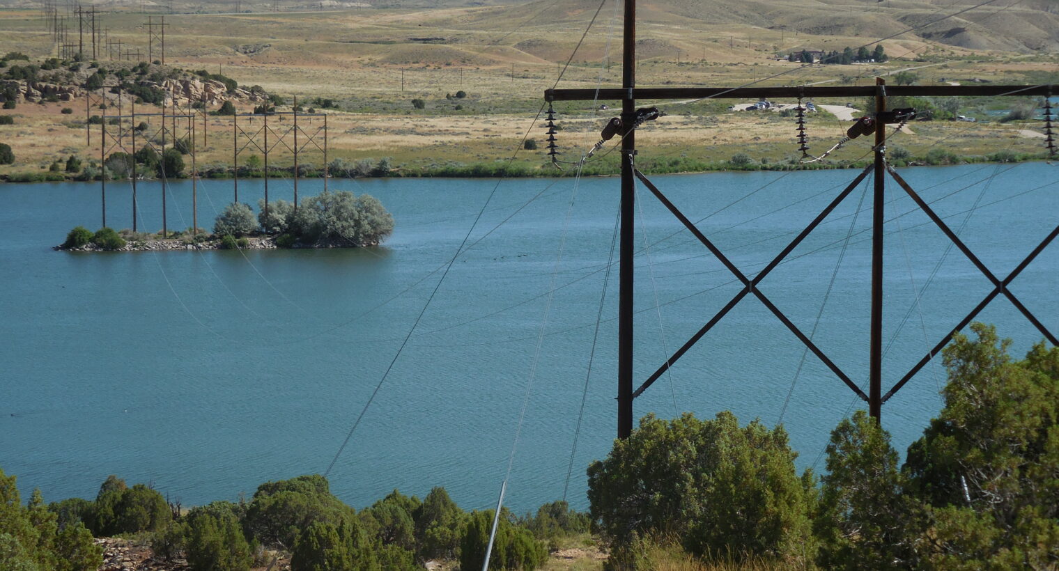 Transmission lines stretching across a lake, supported by tall wooden towers with insulators. In the background, rolling hills and sparse vegetation extend under a clear blue sky.