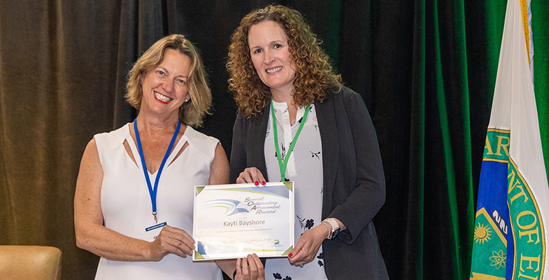 Two women are smiling and holding an award certificate, with a backdrop of a stage and the Department of Energy flag visible.
