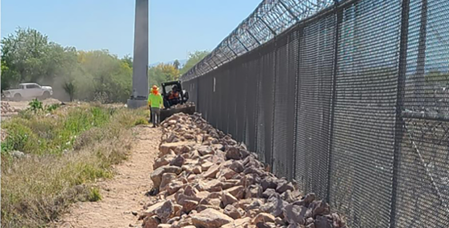 Photo: Counstruction workers use skid-steer to place large angular boulders along chain-link fence.