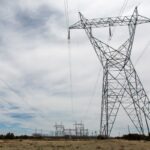 Image of a steel-lattice transmission tower and power lines in a desert landscape with an electrical substation in the distance.