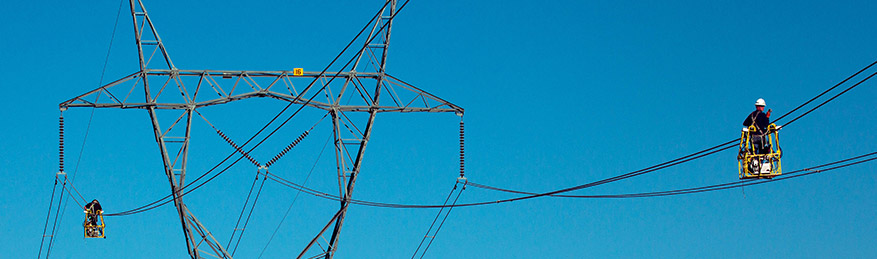 Two line-workers traversing powerlines on spacer carts to replace powerline spacers.