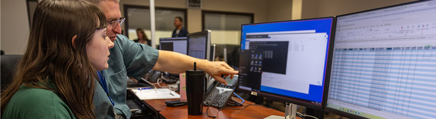 Female graduate learning on the job in front of multiple monitors with a more experienced employee pointing out details.